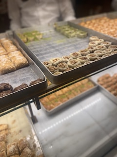 Several trays of various types of desserts are on display, including pastries with visible nuts and powdered sugar. The background shows more trays of similar desserts, creating a vibrant and appetizing arrangement.