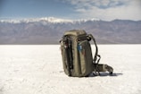 A backpack is placed on an expansive white salt flat with a mountain range in the background. The sky is partly cloudy, adding depth to the landscape. The backpack is equipped with a carabiner and other gear, suggesting a readiness for adventure or travel.