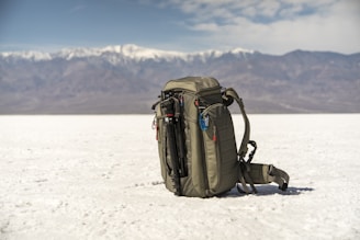 A backpack is placed on an expansive white salt flat with a mountain range in the background. The sky is partly cloudy, adding depth to the landscape. The backpack is equipped with a carabiner and other gear, suggesting a readiness for adventure or travel.