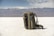 A backpack is placed on an expansive white salt flat with a mountain range in the background. The sky is partly cloudy, adding depth to the landscape. The backpack is equipped with a carabiner and other gear, suggesting a readiness for adventure or travel.