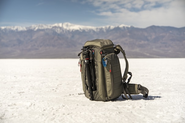 A backpack is placed on an expansive white salt flat with a mountain range in the background. The sky is partly cloudy, adding depth to the landscape. The backpack is equipped with a carabiner and other gear, suggesting a readiness for adventure or travel.