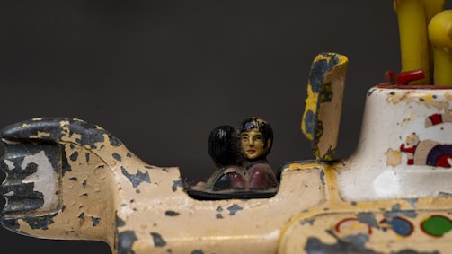 A close-up view of a vintage, worn-out toy featuring a small, painted figurine of a person with detailed facial features. The toy is primarily beige and has areas with chipped paint exposing darker layers underneath. There is a small, faded illustration of a Santa Claus and stars to the right.