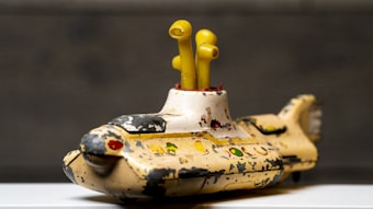 A vintage toy submarine with a worn and chipped paint finish, featuring vibrant yellow periscopes. The submarine's exterior is primarily yellow with decorative red, green, and black accents, placed against a neutral background.