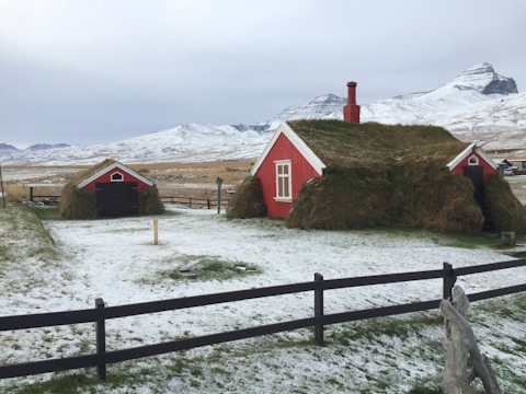 A rural scene features two turf-covered houses with red facades, set against a backdrop of snow-covered fields and mountains. The landscape is open and expansive, with a wooden fence delineating the property. The sky is overcast, casting a cold and serene atmosphere over the area.