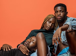 Three individuals are posed closely together against a solid orange background. One person with braided hair is sitting in the center, flanked by two other individuals, one lying down and the other sitting with a relaxed demeanor. The expressions are calm and introspective.