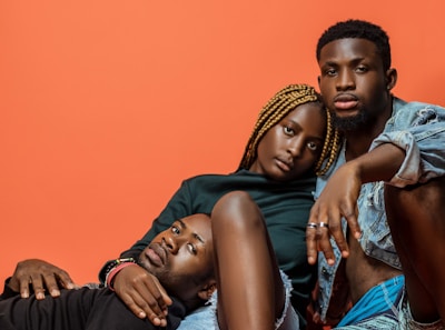 Three individuals are posed closely together against a solid orange background. One person with braided hair is sitting in the center, flanked by two other individuals, one lying down and the other sitting with a relaxed demeanor. The expressions are calm and introspective.