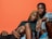 Three individuals are posed closely together against a solid orange background. One person with braided hair is sitting in the center, flanked by two other individuals, one lying down and the other sitting with a relaxed demeanor. The expressions are calm and introspective.