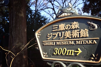 A sign indicating directions to the Ghibli Museum, Mitaka. The sign features a large Totoro figure at the top and displays text in both Japanese and English. An arrow points to the right, directing visitors to the museum 300 meters ahead. The sign is adorned with small leaf details and set against a background of trees.