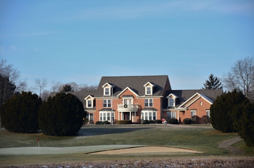 A large, red-brick house with white trim and multiple windows, situated in a rural setting. The house is surrounded by a well-manicured lawn with some trees. In the foreground, there is a golf course green with a sand bunker and a flag.