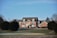 A large, red-brick house with white trim and multiple windows, situated in a rural setting. The house is surrounded by a well-manicured lawn with some trees. In the foreground, there is a golf course green with a sand bunker and a flag.