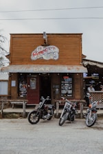 A rustic wooden building serves as a cowboy-themed bar with a vintage sign above the entrance. Three motorcycles are parked in front of the bar, contributing to the rugged and adventurous atmosphere. A man can be seen sitting at a counter inside the bar, adding to the lively and social vibe.
