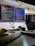 A computer workstation featuring two monitors. The left monitor displays code with a dark theme, surrounded by multiple open tabs and code editors. The right monitor has a dark-themed interface showing a file directory structure. Below the screens, there is a keyboard, books, a pen, and a pair of black headphones. A small decorative globe and a stack of books are situated beneath one of the screens, while sticky notes are placed above the monitors.