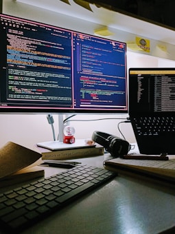 A computer workstation featuring two monitors. The left monitor displays code with a dark theme, surrounded by multiple open tabs and code editors. The right monitor has a dark-themed interface showing a file directory structure. Below the screens, there is a keyboard, books, a pen, and a pair of black headphones. A small decorative globe and a stack of books are situated beneath one of the screens, while sticky notes are placed above the monitors.