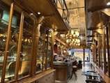 A cozy and elegant interior of a café or restaurant with ornate wooden cabinetry and shelving along the walls filled with glassware and decorative items. The space is well-lit with warm lighting from multiple chandelier-style fixtures. A long counter with bar stools is visible, and there is a person working behind the counter. The floor is patterned, adding to the classic and sophisticated atmosphere.