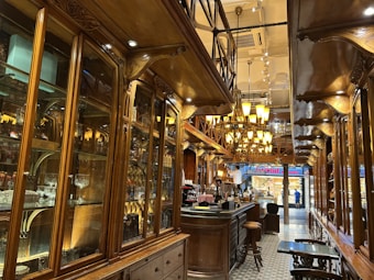 A cozy and elegant interior of a café or restaurant with ornate wooden cabinetry and shelving along the walls filled with glassware and decorative items. The space is well-lit with warm lighting from multiple chandelier-style fixtures. A long counter with bar stools is visible, and there is a person working behind the counter. The floor is patterned, adding to the classic and sophisticated atmosphere.
