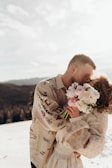 A couple is embracing and kissing in a snowy landscape. The man is wearing a patterned shirt with intricate designs, while the woman holds a bouquet of pink and white flowers. The sun is bright, casting a warm glow over the scene and softening the details of the background, which shows a view of distant hills.