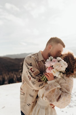 A couple is embracing and kissing in a snowy landscape. The man is wearing a patterned shirt with intricate designs, while the woman holds a bouquet of pink and white flowers. The sun is bright, casting a warm glow over the scene and softening the details of the background, which shows a view of distant hills.