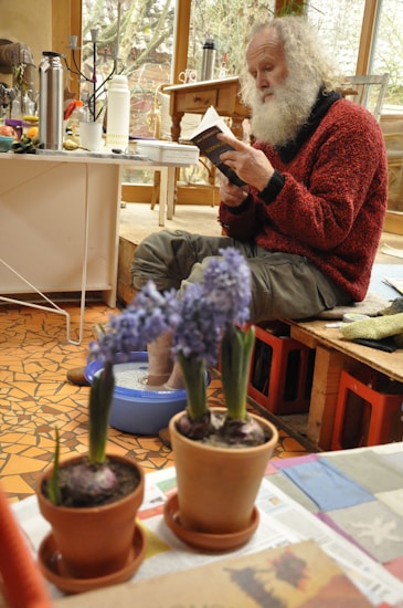An elderly man with a white beard is sitting indoors, engrossed in a book while soaking his feet in a blue basin. The environment is warm and cozy, with potted purple flowers on a table in the foreground. Various household items, including a thermos and bottles, are scattered around, giving a lived-in feel.