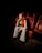 A woman with long red hair and wearing striped black-and-white pants is seated in a dimly lit theater. The seats around her are red and black, and she is positioned thoughtfully with her hand near her face.