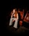 A woman with long red hair and wearing striped black-and-white pants is seated in a dimly lit theater. The seats around her are red and black, and she is positioned thoughtfully with her hand near her face.