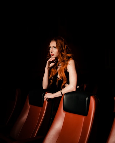 A woman with long red hair is sitting in a dimly lit theater, leaning forward with her chin resting on her hand. She wears a black dress and gazes thoughtfully into the distance, surrounded by red theater seats.