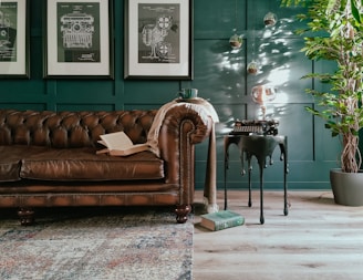 A cozy and stylish living room setup featuring a classic brown leather Chesterfield sofa adorned with an open book and a throw blanket. Three framed technical drawings hang on the dark teal wall. A side table with a vintage typewriter and a unique light bulb adds character. A leafy potted plant sits in the corner, with a textured rug partially covering the wooden floor.