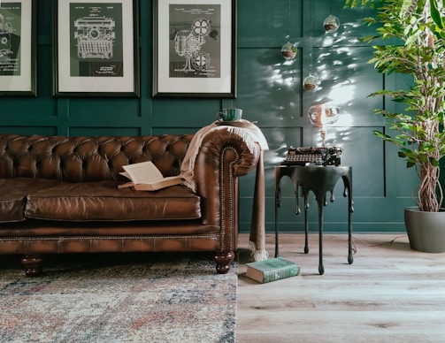 A cozy and stylish living room setup featuring a classic brown leather Chesterfield sofa adorned with an open book and a throw blanket. Three framed technical drawings hang on the dark teal wall. A side table with a vintage typewriter and a unique light bulb adds character. A leafy potted plant sits in the corner, with a textured rug partially covering the wooden floor.