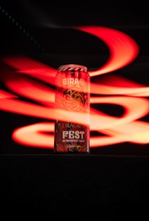 A can of Bira 91 beer is prominently displayed against a black background, with dynamic red light streaks swirling around it. The can features branding and symbols including a dragon and the word 'FEST' in bold lettering, indicating an Oktoberfest Lager edition.