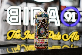 A colorful beer can with a vibrant design featuring an assortment of playful patterns and fireworks. The can displays the brand 'Bira 91' prominently, and the background showcases a neon sign with the brand logo and name, adding brightness to the scene.