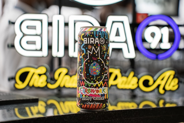 A colorful beer can with a vibrant design featuring an assortment of playful patterns and fireworks. The can displays the brand 'Bira 91' prominently, and the background showcases a neon sign with the brand logo and name, adding brightness to the scene.