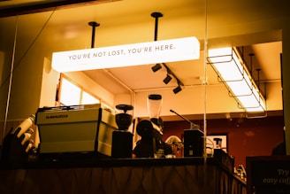 A warmly lit coffee shop interior featuring a counter with a large espresso machine and a coffee grinder. Behind the counter, there are baristas preparing drinks. The ceiling showcases modern light fixtures, and a prominent lightbox above displays the words 'YOU'RE NOT LOST, YOU'RE HERE.'