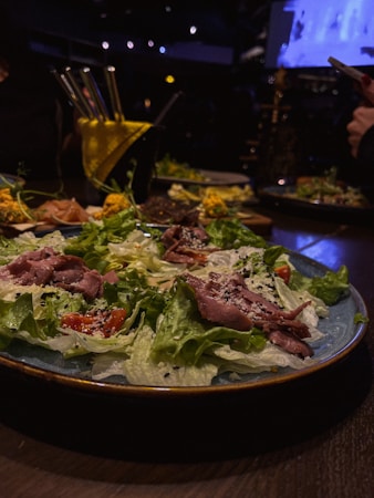 A plate of fresh salad with slices of cooked meat, lettuce, cherry tomatoes, and sprinkled sesame seeds. The setting includes dim lighting typical of a cozy restaurant environment. Bright yellow straws emerge from a drink in the background.