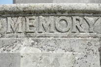 Carved stone surface with the word 'MEMORY' prominently etched in large, bold letters. The stone exhibits weathering and mossy growth, suggesting it has been exposed to the elements for an extended period of time.