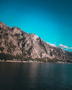 A vast mountain range rises steeply from the edge of a calm body of water, under a clear, bright blue sky. The rocky surface of the mountain is interspersed with patches of greenery. Small clusters of buildings or houses can be seen at the base of the mountains, suggesting a settlement along the shoreline.