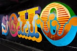 A vibrant and colorful graffiti mural on a dark wall, featuring large stylized letters with swirling patterns and a mix of bright hues including yellow, pink, orange, blue, and green. The artwork's bold design is illuminated against a shadowy urban background.