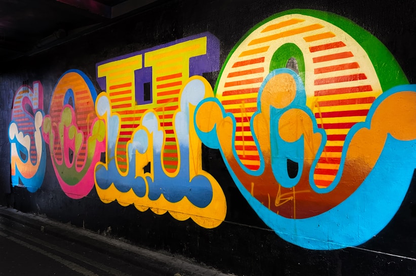 A vibrant and colorful graffiti mural on a dark wall, featuring large stylized letters with swirling patterns and a mix of bright hues including yellow, pink, orange, blue, and green. The artwork's bold design is illuminated against a shadowy urban background.