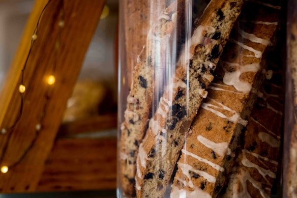 A close-up view of a glass jar filled with biscotti cookies, which are drizzled with white icing and have visible chocolate chips. The jar is positioned next to rustic wooden beams decorated with small string lights.