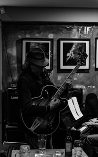 A musician wearing a fedora plays a guitar passionately in a cozy indoor setting. The environment features framed photographs on the wall and a piano with sheet music, while glasses and a bottle rest on a table in the foreground.