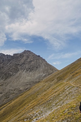 A rugged mountain peak rises against a partly cloudy blue sky. The slopes are covered in a mix of rocky terrain and patches of grass. A narrow path winds up the side, suggesting a hiking trail.