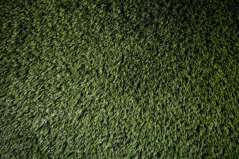 The image shows a close-up view of a carpet made from artificial grass, exhibiting a vibrant shade of green. The carpet is densely packed with numerous individual blades of grass, creating an intricate pattern that adds depth and texture to the scene. The background appears to be a light gray color, providing a neutral backdrop that allows the green carpet to stand out prominently.