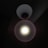 A single bright circular light source is centered within a dark background. The light appears to be an LED bulb encased in a white lampshade that directs the light downwards, creating a halo effect around the light.