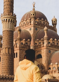 An elaborate mosque with intricate architectural details and a prominent minaret stands in sunlight. A person in a yellow jacket is gazing at the structure, highlighting the spiritual and cultural ambiance.