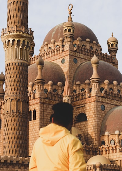 An elaborate mosque with intricate architectural details and a prominent minaret stands in sunlight. A person in a yellow jacket is gazing at the structure, highlighting the spiritual and cultural ambiance.