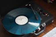 A vintage record player is seen from above, spinning a translucent blue vinyl record labeled 'Midnights'. The player, with its retro design, is placed on a wooden surface. The tonearm is in contact with the record, and small indicator lights are visible on the player.