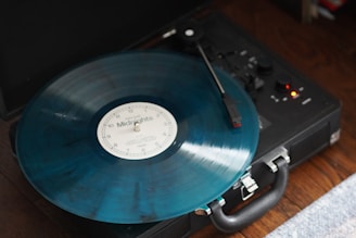 A vintage record player is seen from above, spinning a translucent blue vinyl record labeled 'Midnights'. The player, with its retro design, is placed on a wooden surface. The tonearm is in contact with the record, and small indicator lights are visible on the player.