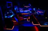 A high-tech music production studio illuminated by neon blue and red lights. Various equipment including keyboards, computer monitors, large speakers, and mixing consoles are present. An ergonomic office chair is positioned in front of the central monitoring station.