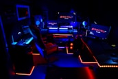 A high-tech music production studio illuminated by neon blue and red lights. Various equipment including keyboards, computer monitors, large speakers, and mixing consoles are present. An ergonomic office chair is positioned in front of the central monitoring station.