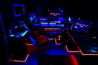 A high-tech music production studio illuminated by neon blue and red lights. Various equipment including keyboards, computer monitors, large speakers, and mixing consoles are present. An ergonomic office chair is positioned in front of the central monitoring station.