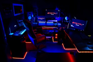 A high-tech music production studio illuminated by neon blue and red lights. Various equipment including keyboards, computer monitors, large speakers, and mixing consoles are present. An ergonomic office chair is positioned in front of the central monitoring station.