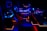 A high-tech music production studio illuminated by neon blue and red lights. Various equipment including keyboards, computer monitors, large speakers, and mixing consoles are present. An ergonomic office chair is positioned in front of the central monitoring station.